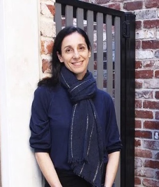 Maria Papas wearing blue shirt and blue scarf, posing by a brick wall, smiling